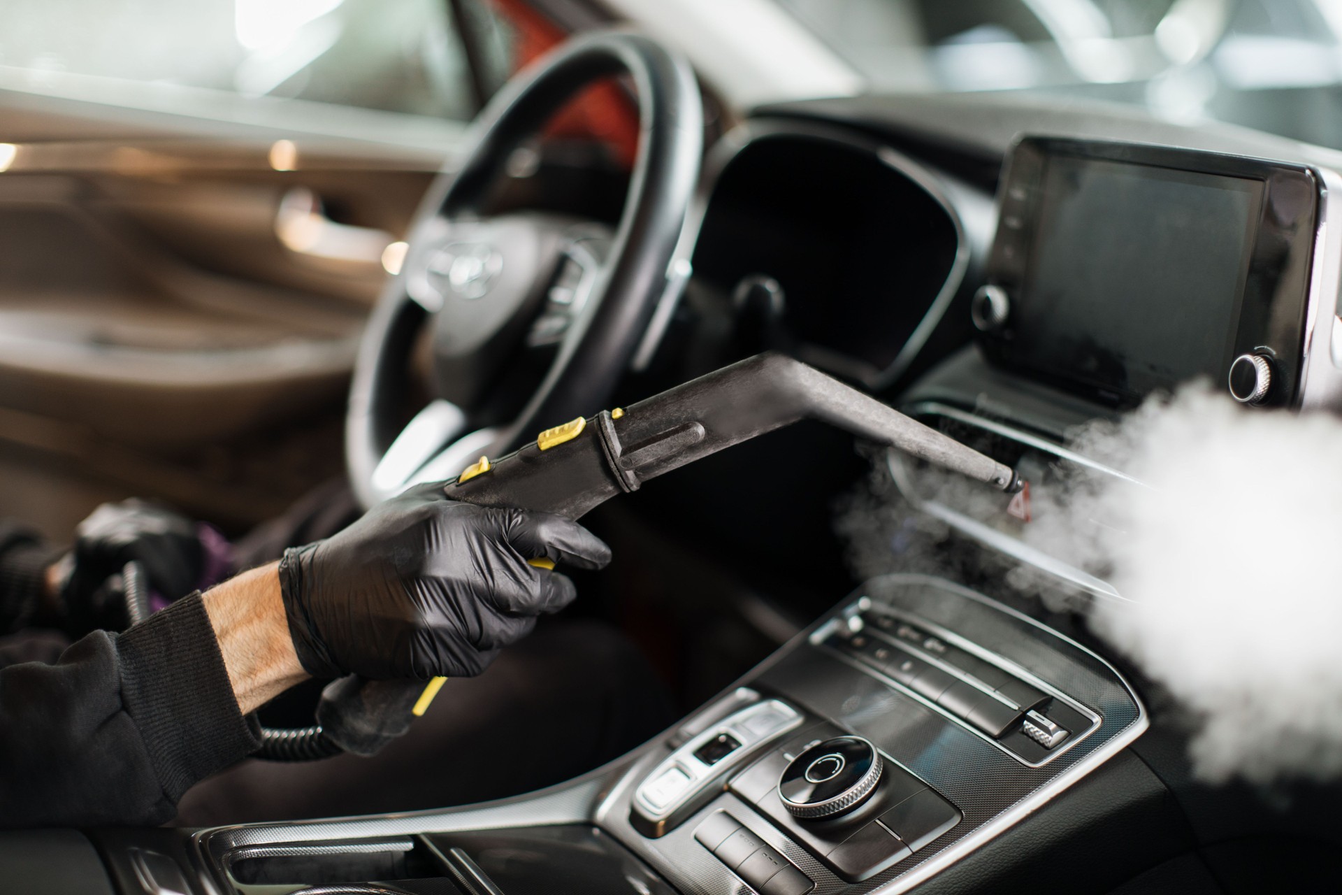 Hands of man in black protective rubber gloves cleaning interior of the car with hot steam cleaner.